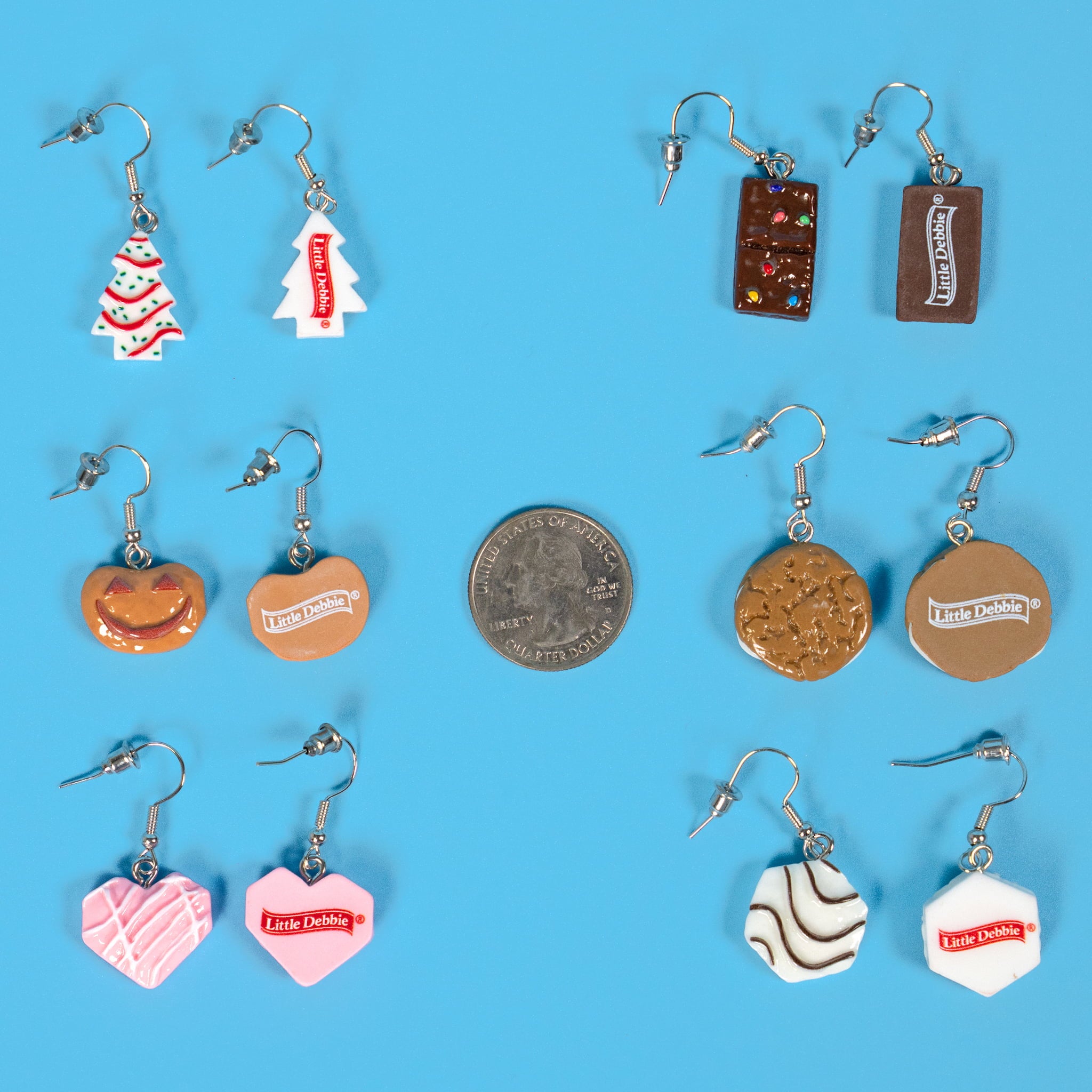 A collection of Little Debbie–inspired earrings displayed on a light blue background, featuring miniature designs of iconic snacks including Christmas Tree Cakes, Cosmic Brownies, Pumpkin Delights, Oatmeal Creme Pies, Strawberry Hearts, and Zebra Cakes. A U.S. quarter is placed in the center for size comparison.