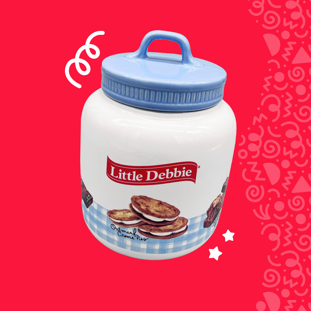 This image features a ceramic Little Debbie cookie jar against a bright red background with playful white doodles. The jar, with a snug-fitting blue lid, is decorated with an image of the Little Debbie logo above a blue gingham banner that wraps around the base. On the banner, appetizing Oatmeal Creme Pies are depicted, adding to the jar's charming appeal. The jar's design conveys a cozy, vintage vibe, ideal for storing and serving treats.