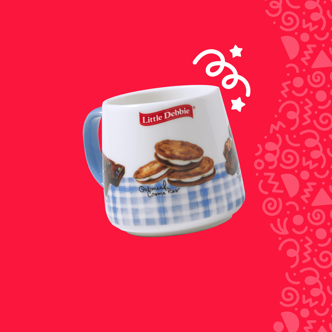 An image of a Little Debbie branded mug on a red background with white swirl patterns. The mug is white with a blue gingham check design around the bottom, featuring an image of stacked Oatmeal Creme Pies and the Little Debbie logo above. The mug has a solid blue handle. The design elements suggest a cozy, home-style kitchen atmosphere.