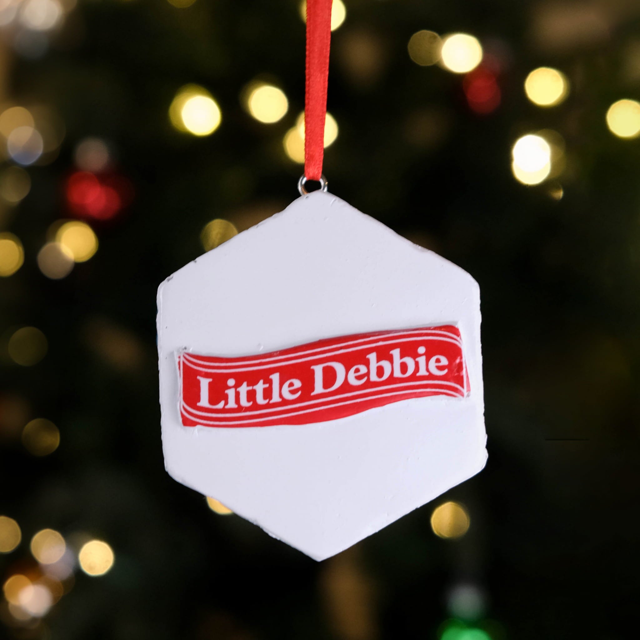 Back of the Little Debbie Zebra Cake Ornament featuring a smooth white surface with the red Little Debbie ribbon logo in the center, hanging by a red ribbon in front of a softly lit Christmas tree.