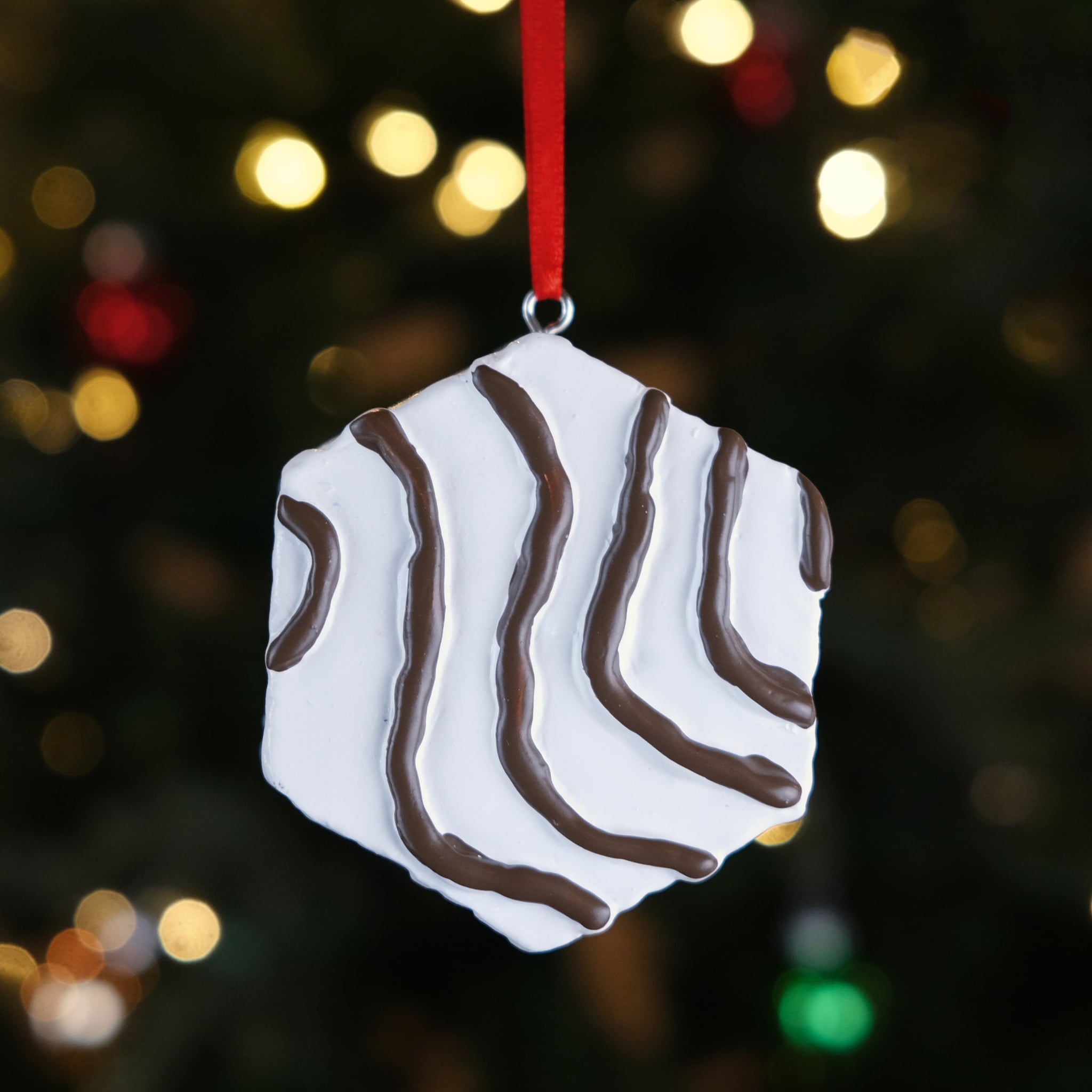 Little Debbie Zebra Cake Ornament featuring a white hexagon-shaped design with wavy chocolate-colored stripes, hanging by a red ribbon in front of a softly lit Christmas tree.